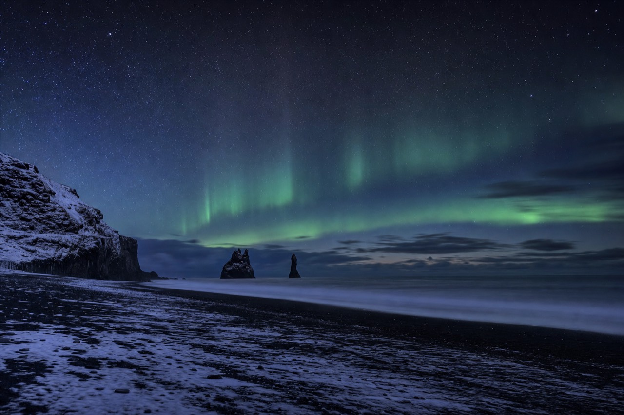 Green northern lights arcing over the sea stacks and black shore at Reynisfjara on a clear winter night